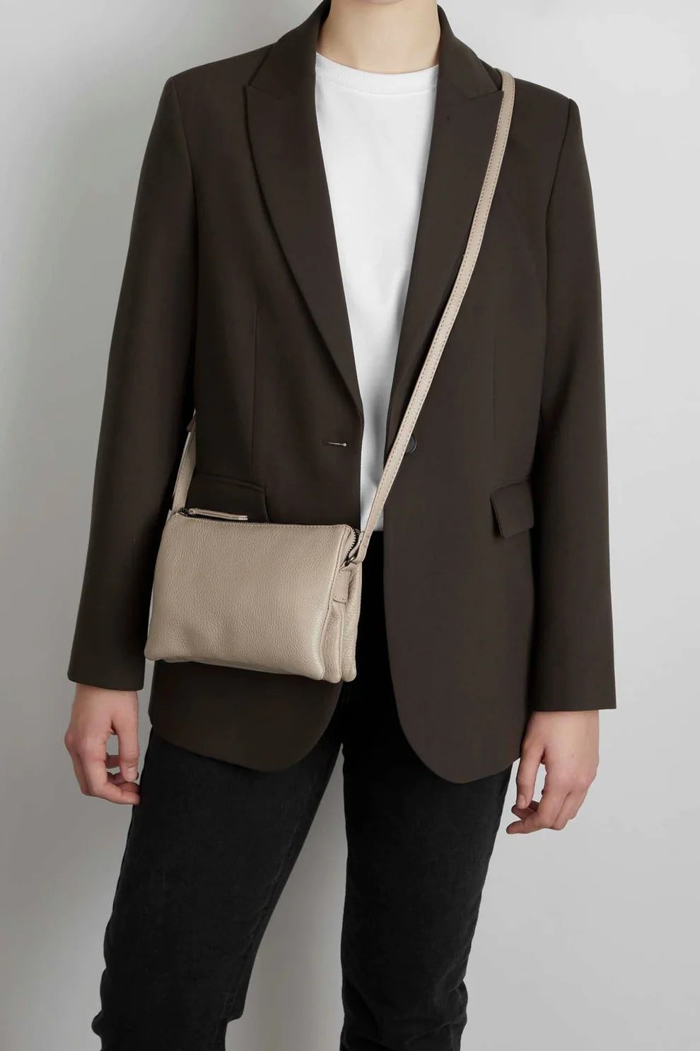 Person wearing a dark blazer and black jeans with a white t-shirt, accessorized with a light beige Markberg Grain Crossbody Bag.