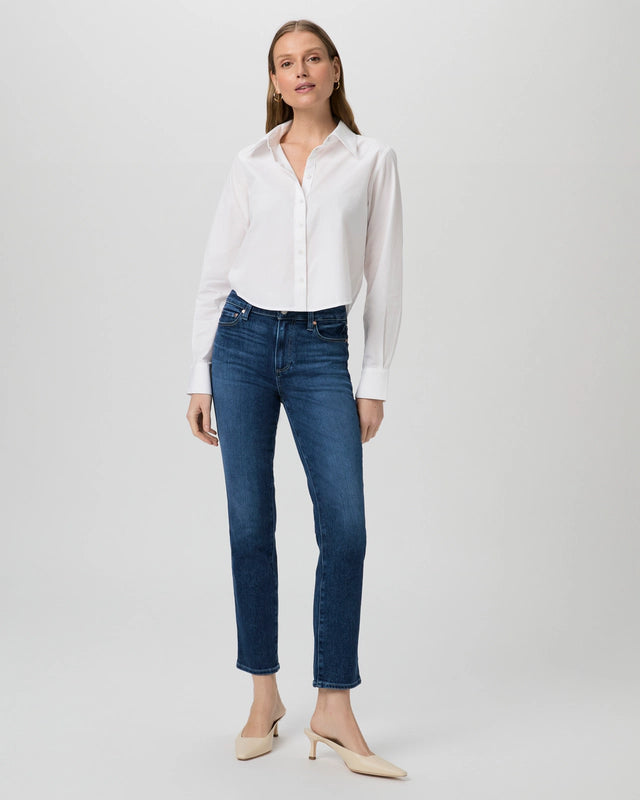 A woman stands against a plain background wearing a white shirt, the Paige Cindy Straight Leg - Kindered jeans with a slim silhouette, and beige heels. Her high-rise denim perfectly complements her sleek ensemble.