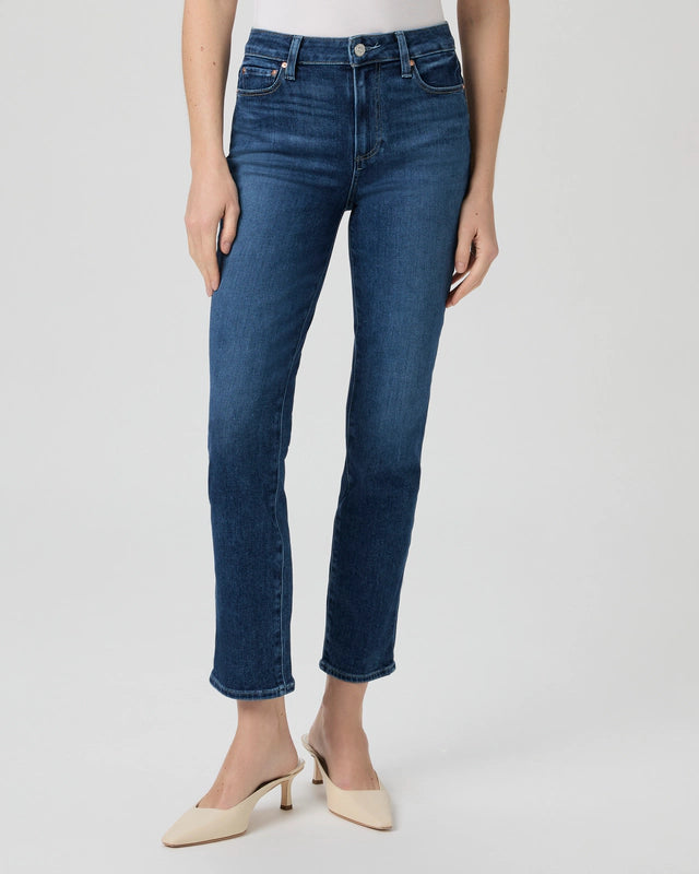 A person models the Cindy Straight Leg - Kindered by Paige against a simple backdrop, highlighting slim leg silhouette dark blue jeans paired with vintage-inspired beige low-heeled shoes.