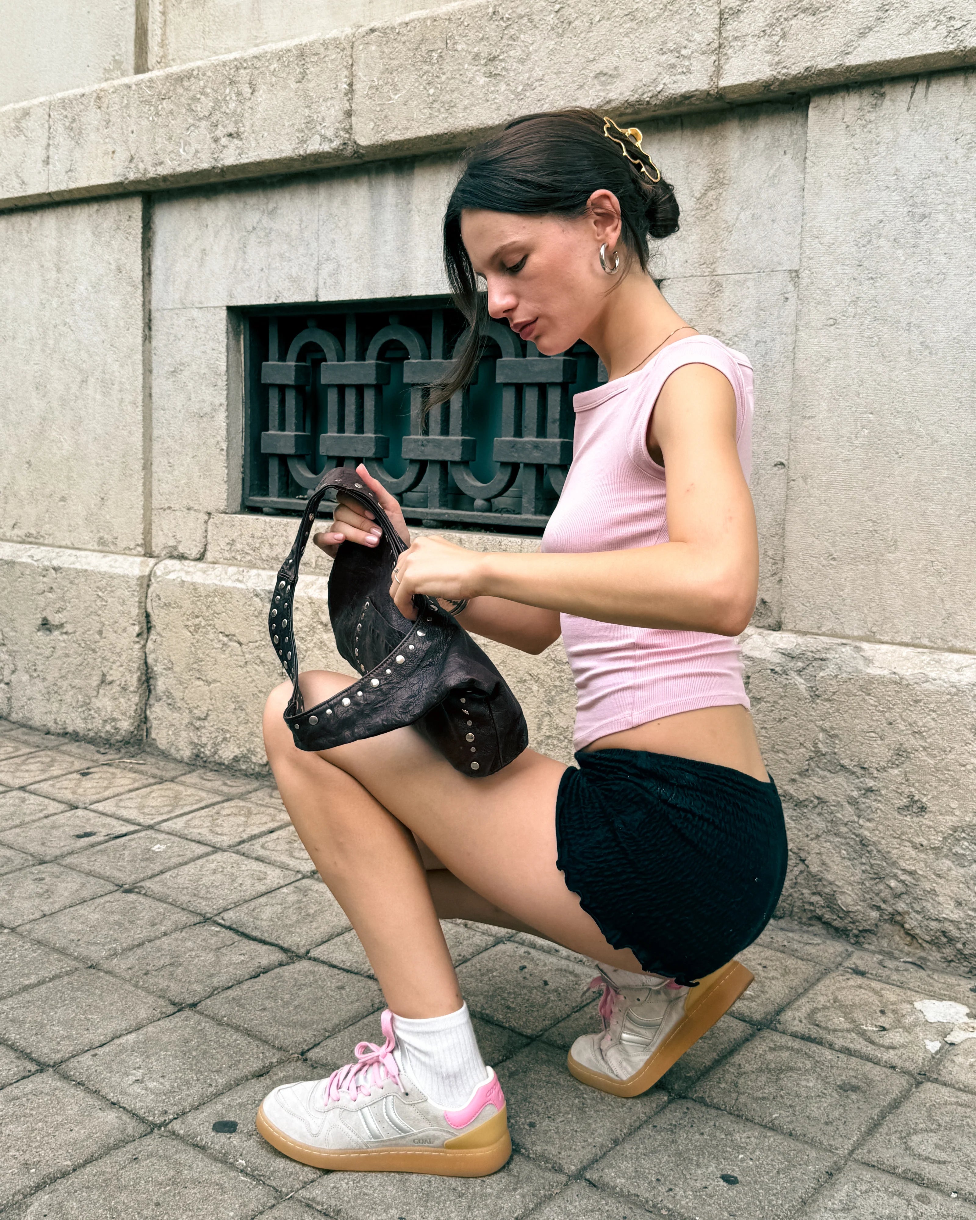 A woman in a pink top and black shorts squats on a city sidewalk, looking into a brown studded handbag while wearing Coolway’s Goal Sneakers in Ice Silver.