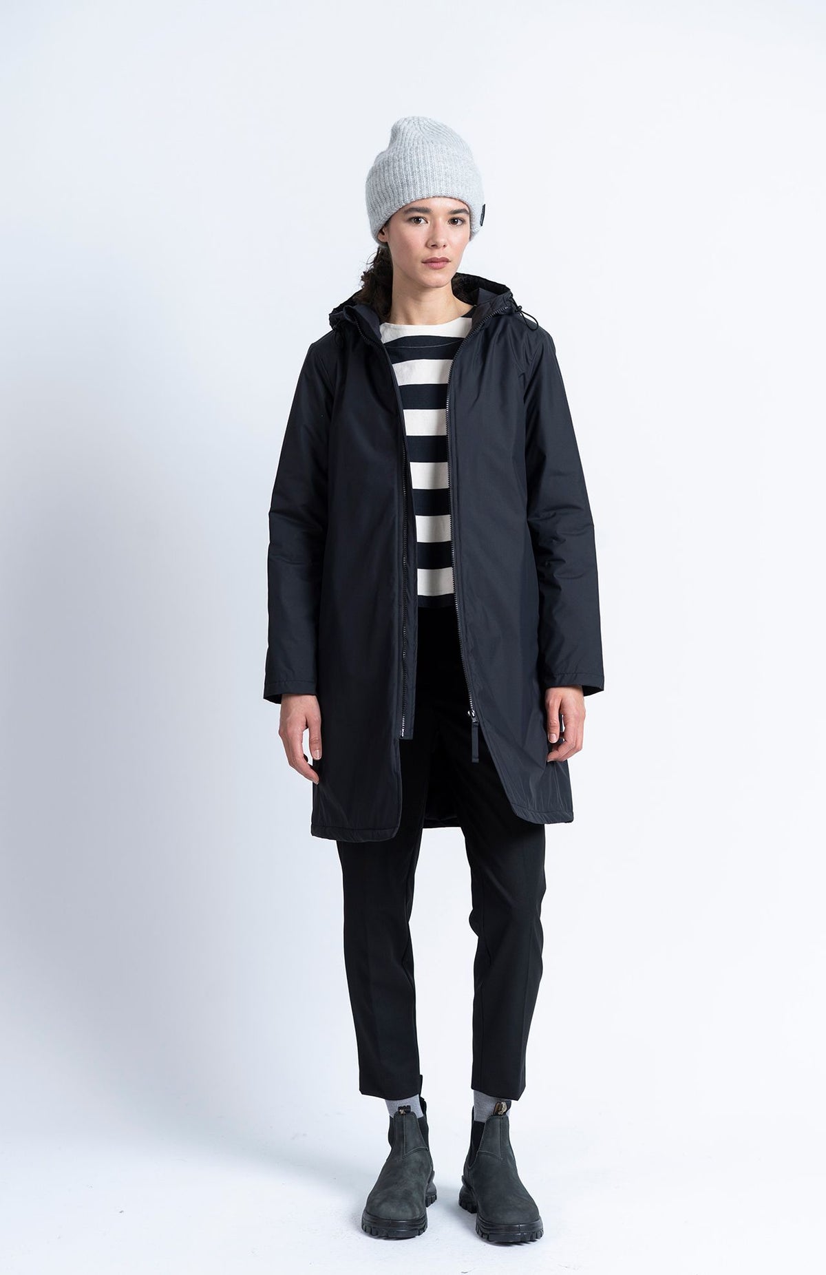 A person wearing a grey beanie, a black hooded parka over a striped shirt, black Alaiak Trouser - Black joggers, and dark ankle boots.