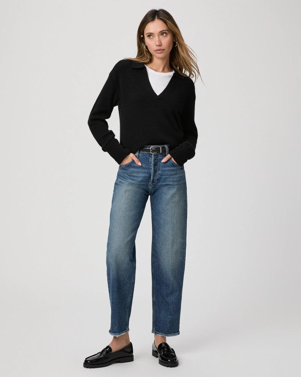 A woman wearing a black sweater, white shirt, and black loafers stands with hands in pockets against a plain background. Her Paige Alexis Barrel Leg jeans in the Lovejoy Distressed medium blue wash add a touch of timeless style to her ensemble.