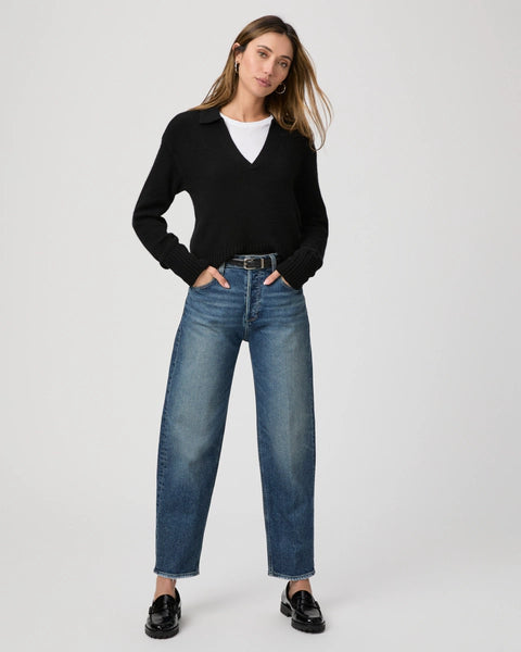 A person wearing a black sweater and white shirt pairs their ensemble with the Paige Alexis Barrel Leg jeans in the Lovejoy Distressed wash, standing against a plain background complemented by sleek black shoes.