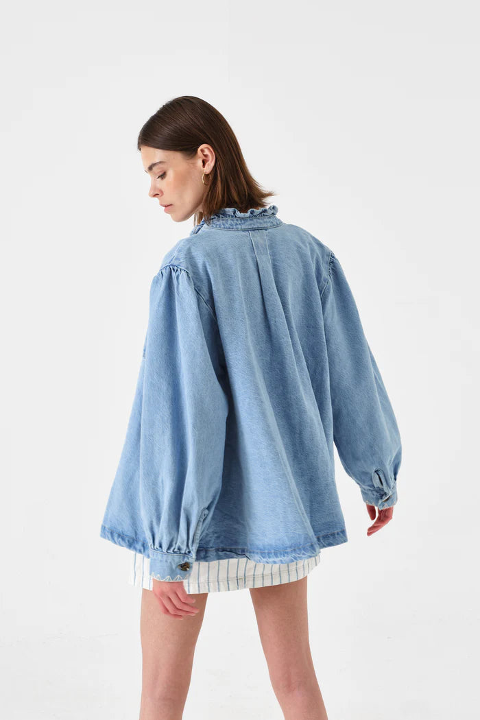 A woman with short brown hair wears the Seventy & Mochi Dana Denim Jacket in Sunbleached, featuring balloon sleeves and a ruffled collar, paired with a white striped skirt. She is facing away.