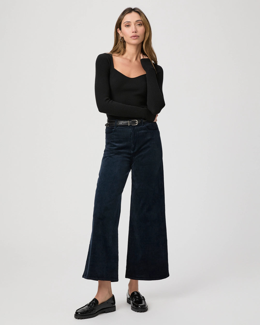 A person stands confidently wearing a black long-sleeve top, Harper Ankle Wide Leg jeans in navy corduroy by Paige, and black loafers against a neutral background.