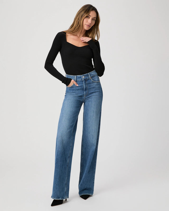 A person models in a Paige Sasha Wide Leg Jean and a black long-sleeve top, with one hand in the pocket, against a plain background.