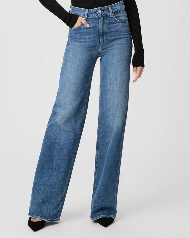 Sporting Paige's Sasha Wide Leg Jean, a vintage high-waisted style, paired with a black long-sleeve top, and standing with one hand in the pocket.