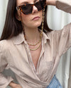 Gold/Tort sunglasses chain by Talis Chains styled by a person wearing an open striped caramel colured shirt and oversized sunglasses, standing in front of a off white coloured curtain. 