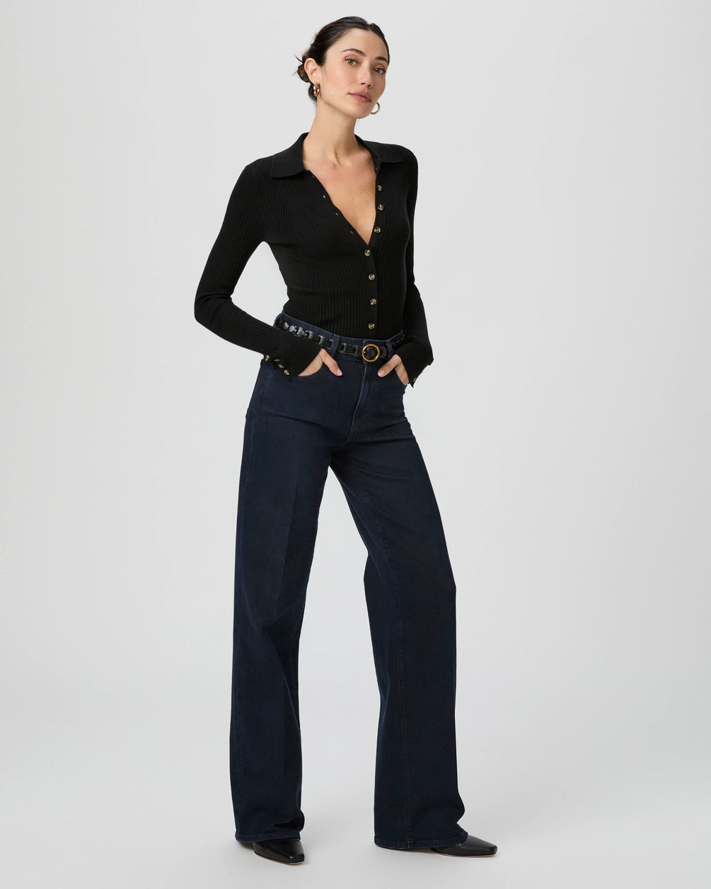 A woman stands against a plain background wearing a black button-up ribbed top, Paige Sasha Wide Leg - Duchess jeans, a black belt, and black shoes, with one hand in her pocket.