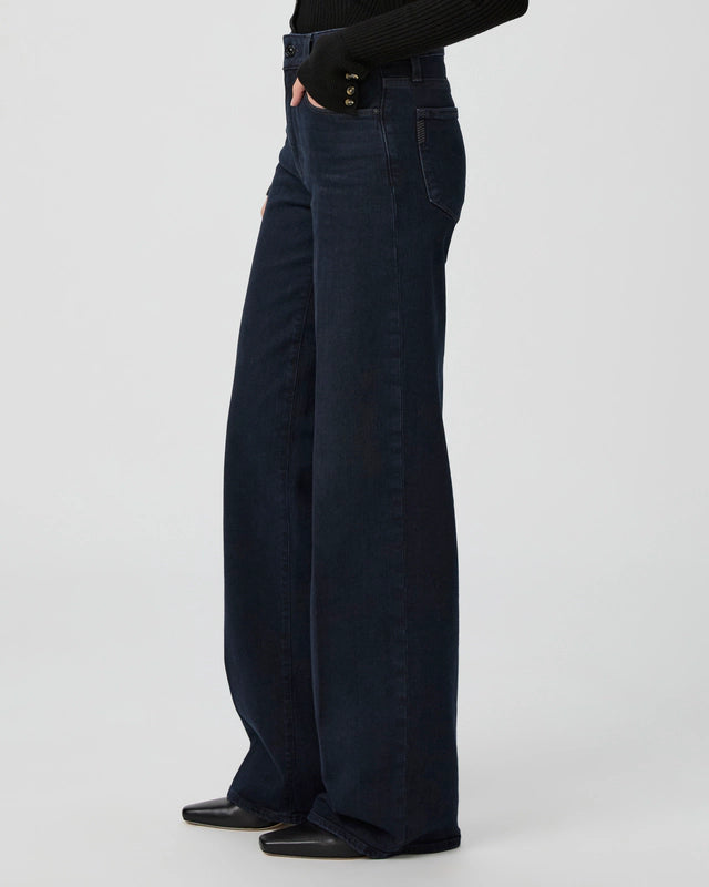 Person wearing high-rise Sasha Wide Leg - Duchess jeans by Paige in a dark wash with black boots, standing with one hand in pocket against a plain light background.