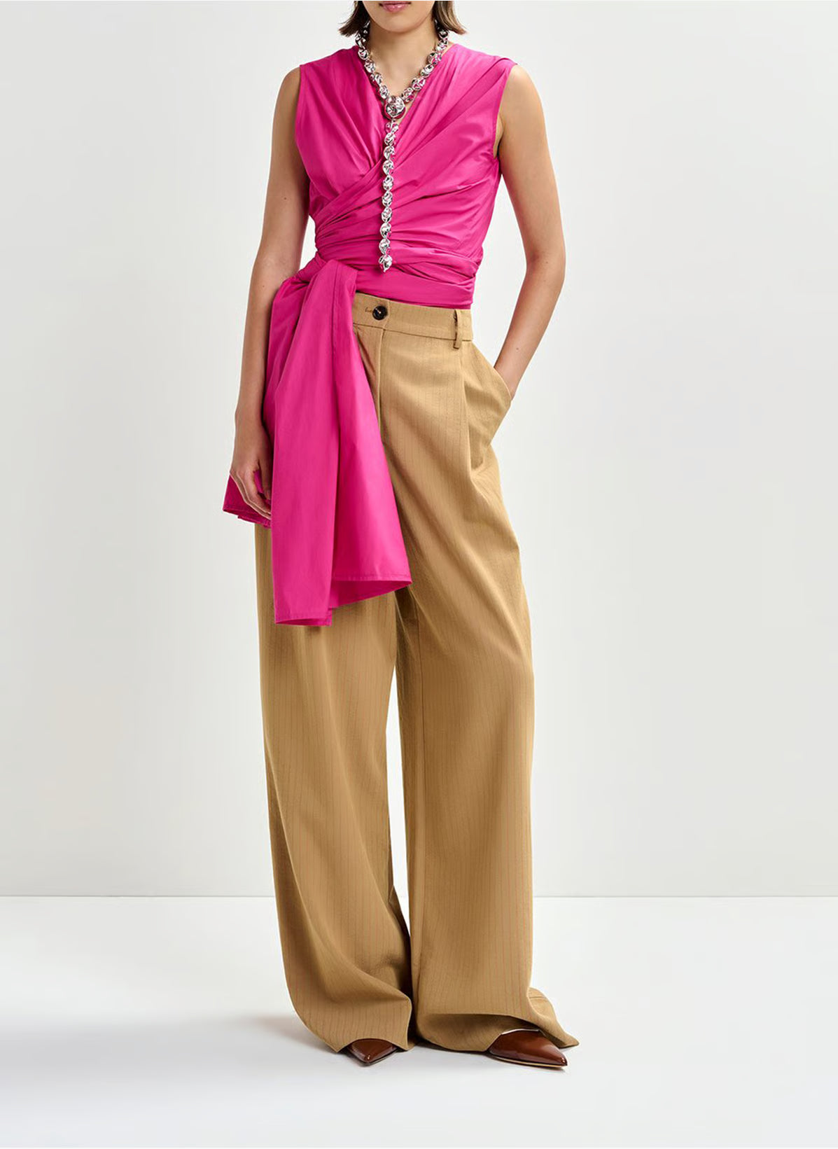 A person in the Essentiel Antwerp Jelani Sleeveless Bow Top in pink, paired with wide-leg, high-waisted beige trousers and brown pointed shoes, stands against a plain white background.