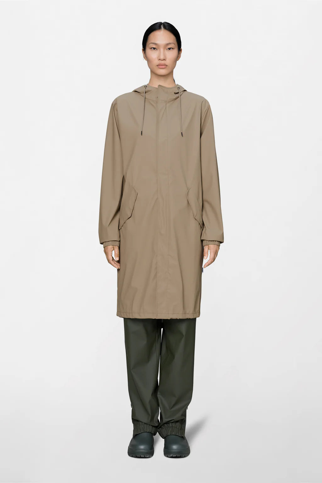 A person stands facing forward against a plain light background, wearing the Rains Fishtail Parka - Core Colours in beige with a hood, paired with dark green pants and black shoes.