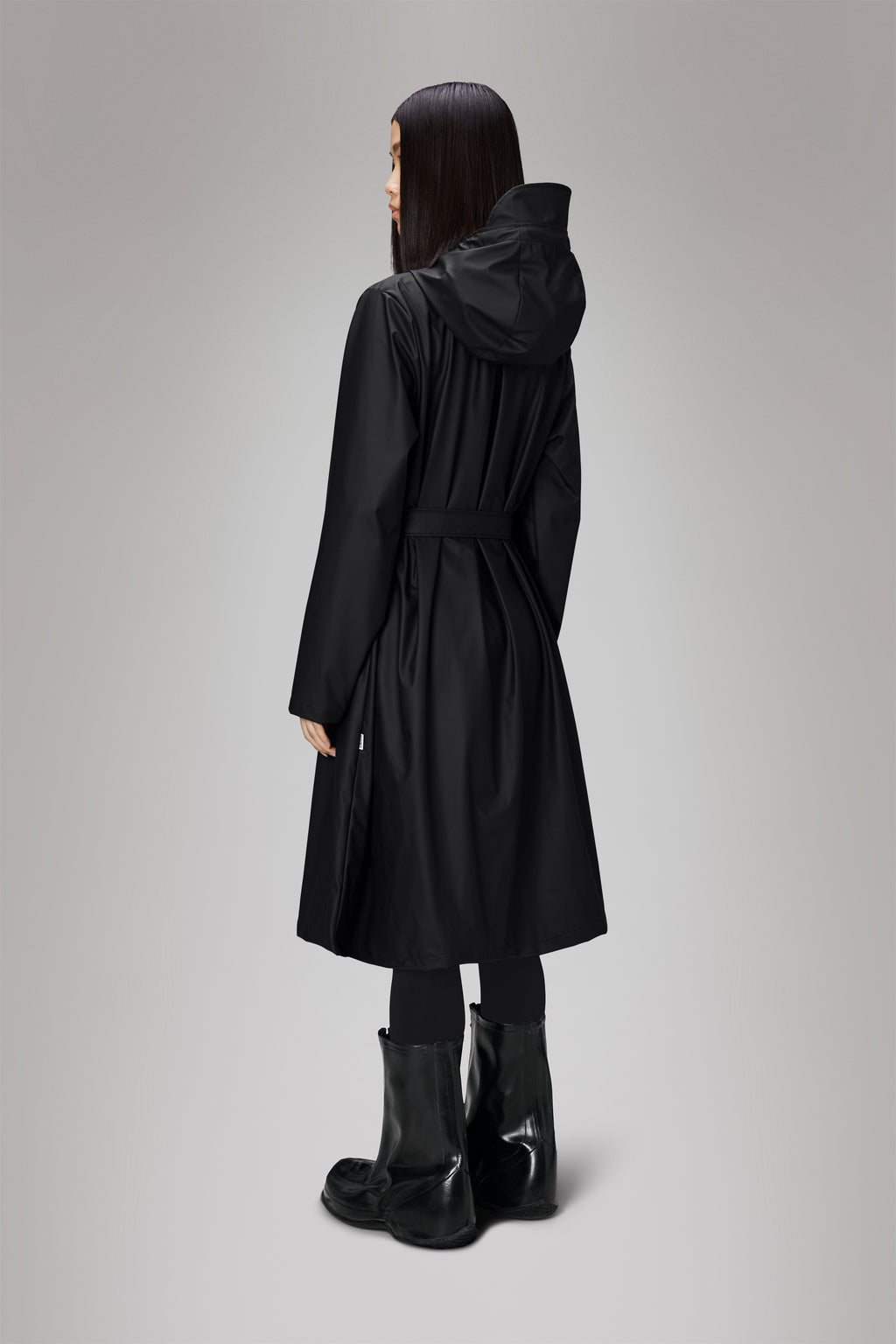 A person stands with their back turned, wearing the A-Line Longer W Jacket from Rains, a stylish long black hooded raincoat in Rains PU fabric for waterproof protection, paired with sturdy black boots.