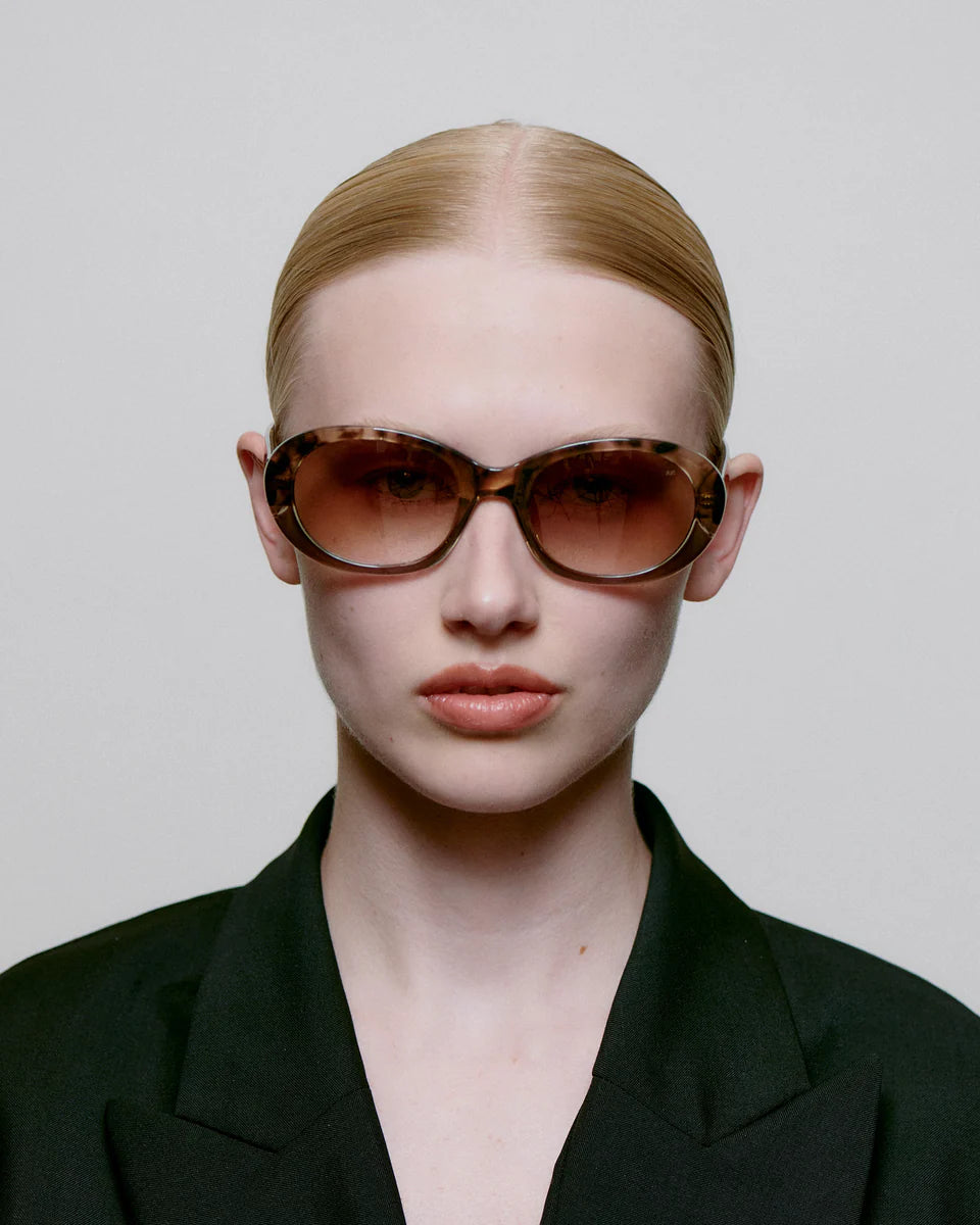 A person with slicked-back blonde hair wears A. Kjærbede's Anma Sunglasses in Coquina / Grey Transparent (UV 400 protection) and a black blazer, facing forward against a plain light background.