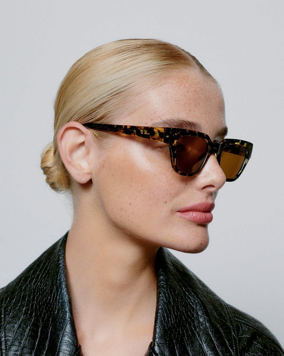 A woman with blonde hair in a low bun models A. Kjærbede's Kaws Sunglasses in Black/Yellow Tortoise, featuring UV 400 protection, paired with a black textured jacket against a plain background.