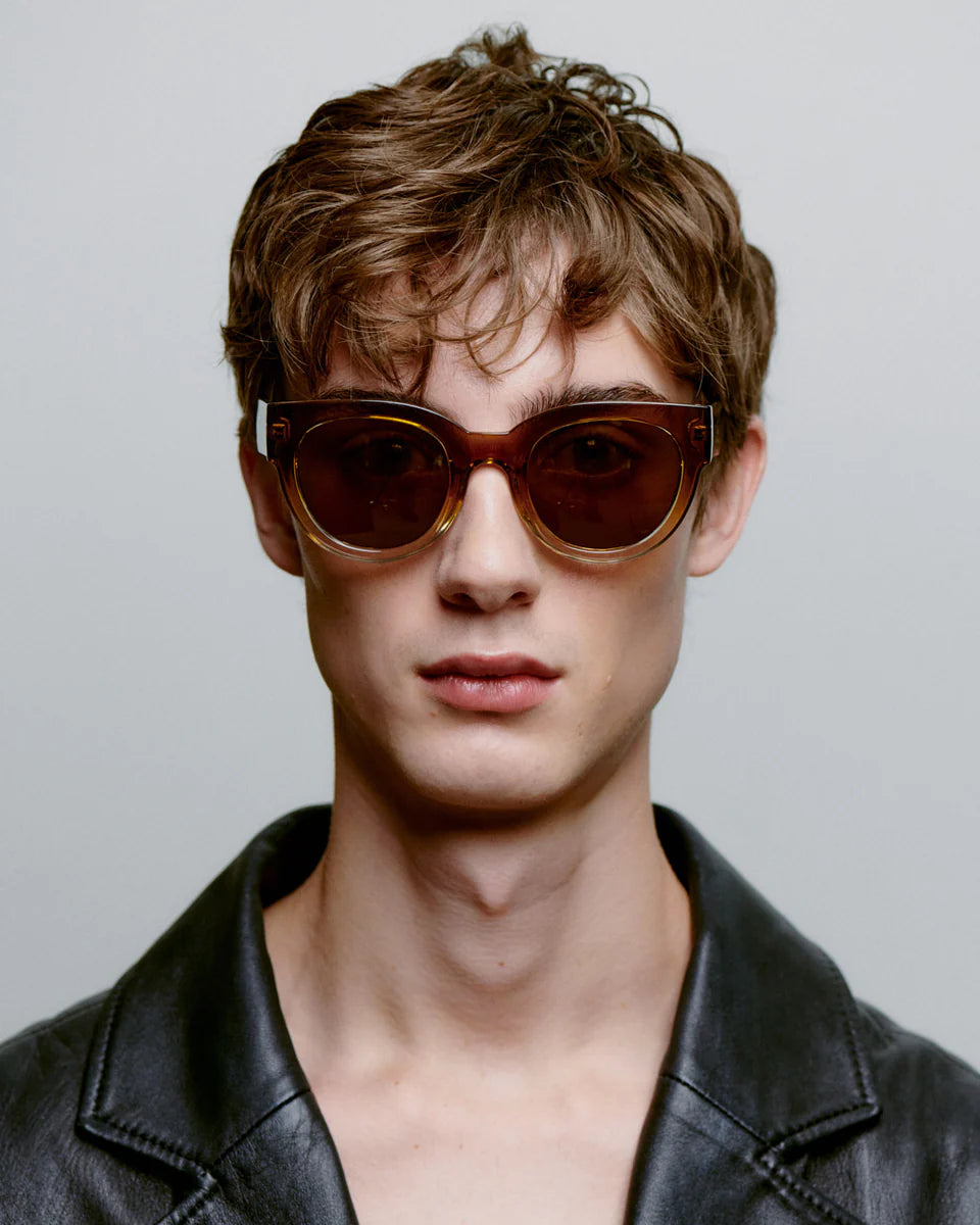 Young man with tousled brown hair wears A. Kjærbede Lilly Sunglasses in Smoke Champagne and a black leather jacket, facing the camera against a plain light gray background.