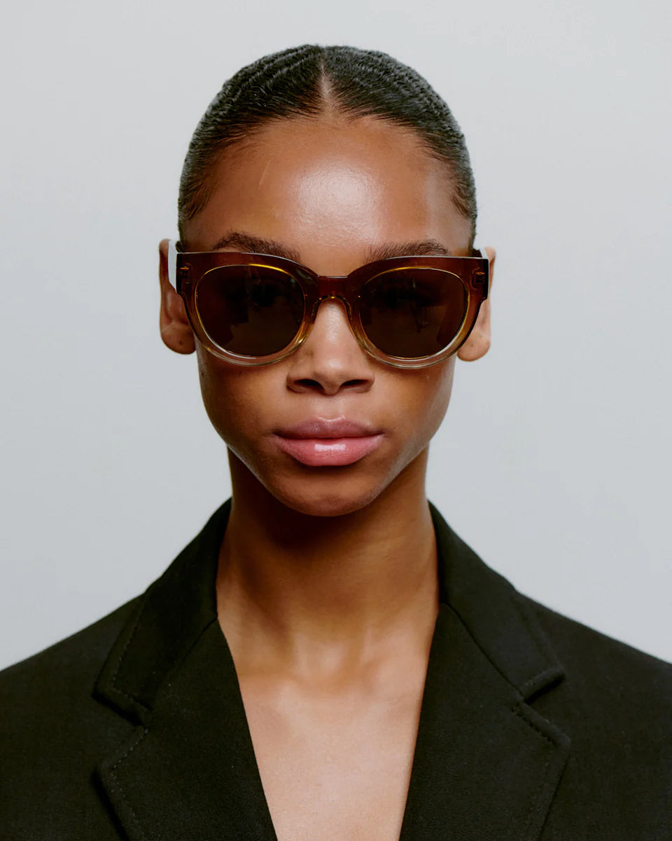 A person with slicked-back hair wears A. Kjærbede's Lilly Sunglasses in Smoke Champagne and a black blazer, facing forward against a plain light background.