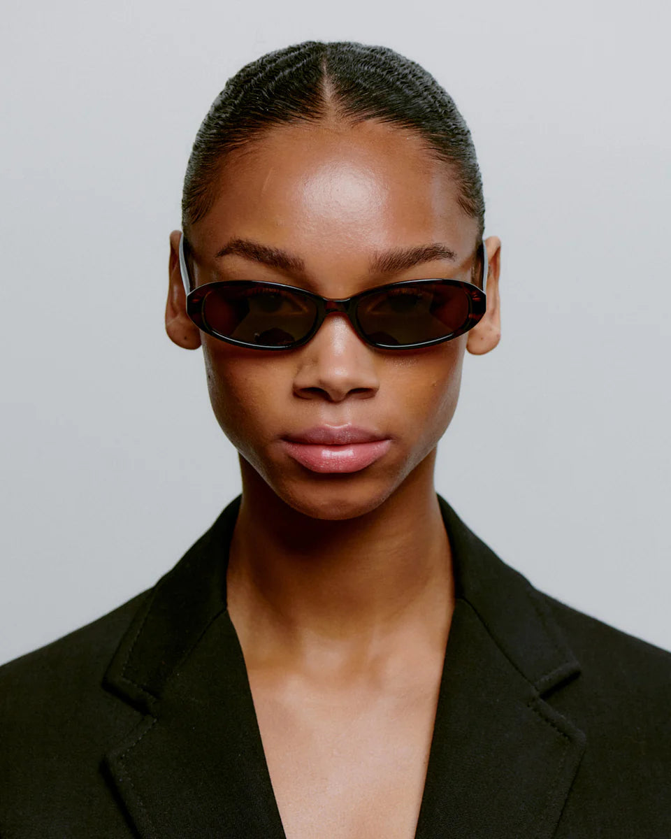 A person with slicked-back hair wears the Macy Sunglasses - Demi Tortoise by A. Kjærbede, featuring UV 400 protection and a narrow frame, paired with a black blazer against a plain light background, embodying sleek Scandinavian style.