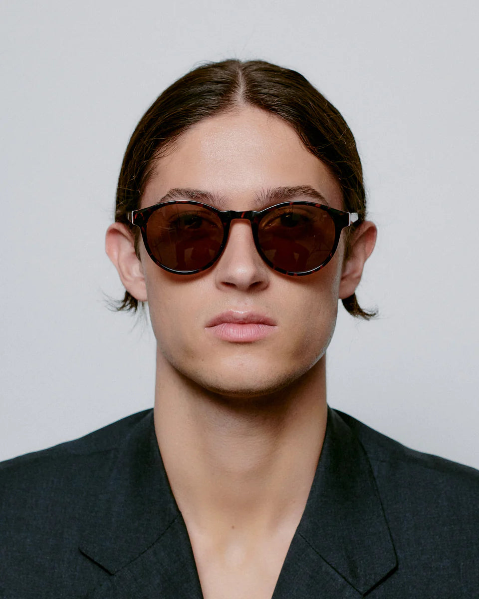 A person with straight brown hair, wearing A. Kjærbede Marvin Sunglasses in Demi Tortoise and a dark blazer, faces the camera against a plain light background.