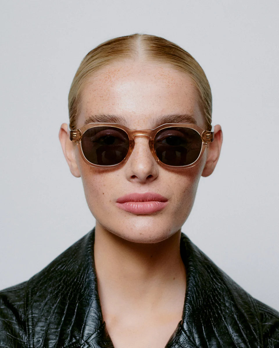 A person with slicked-back blonde hair wears A. Kjærbede Zan Sunglasses in Champagne and a black textured jacket, facing forward against a plain light background.