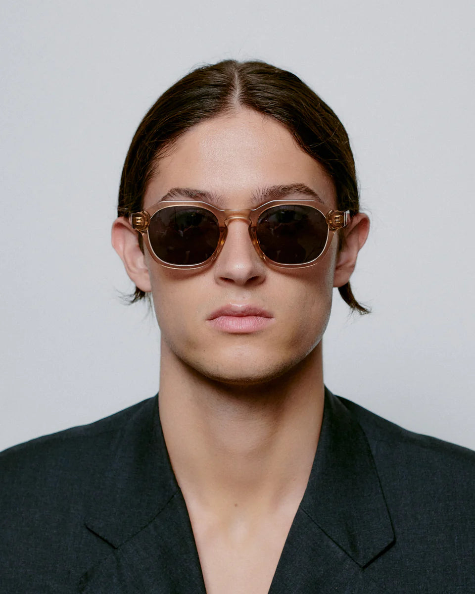 A person with straight brown hair, facing forward against a plain light background, wears A. Kjærbede Zan Sunglasses in Champagne featuring eco-friendly materials and UV 400 protection, paired with a dark blazer.