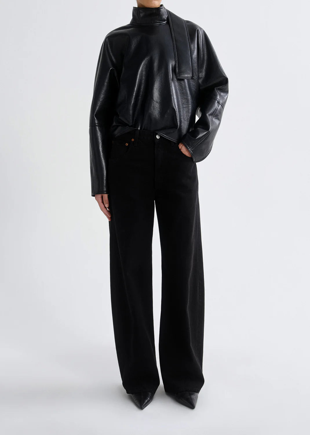 Person wearing a black leather long-sleeve top with neck tie detail, AGOLDE Low Curve Jean - Black, and black pointed-toe shoes stands against a plain light background.