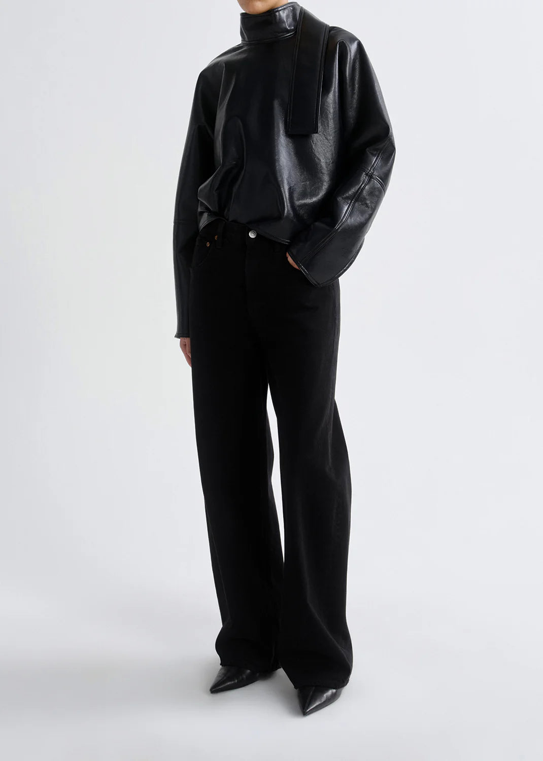 A person wearing AGOLDE's Low Curve Jean - Black, crafted from regenerative cotton, pairs it with a black leather high-neck long-sleeve top and black pointed-toe shoes against a plain light background.