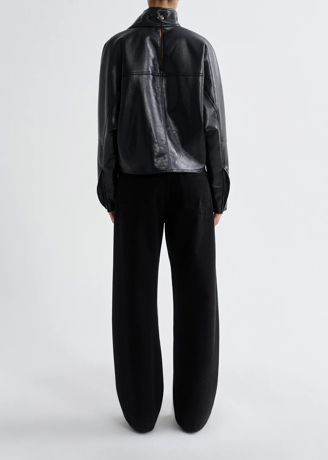 A person faces away from the camera wearing a black leather jacket and AGOLDE’s Low Curve Jean - Black, crafted from regenerative cotton denim, set against a plain light background.