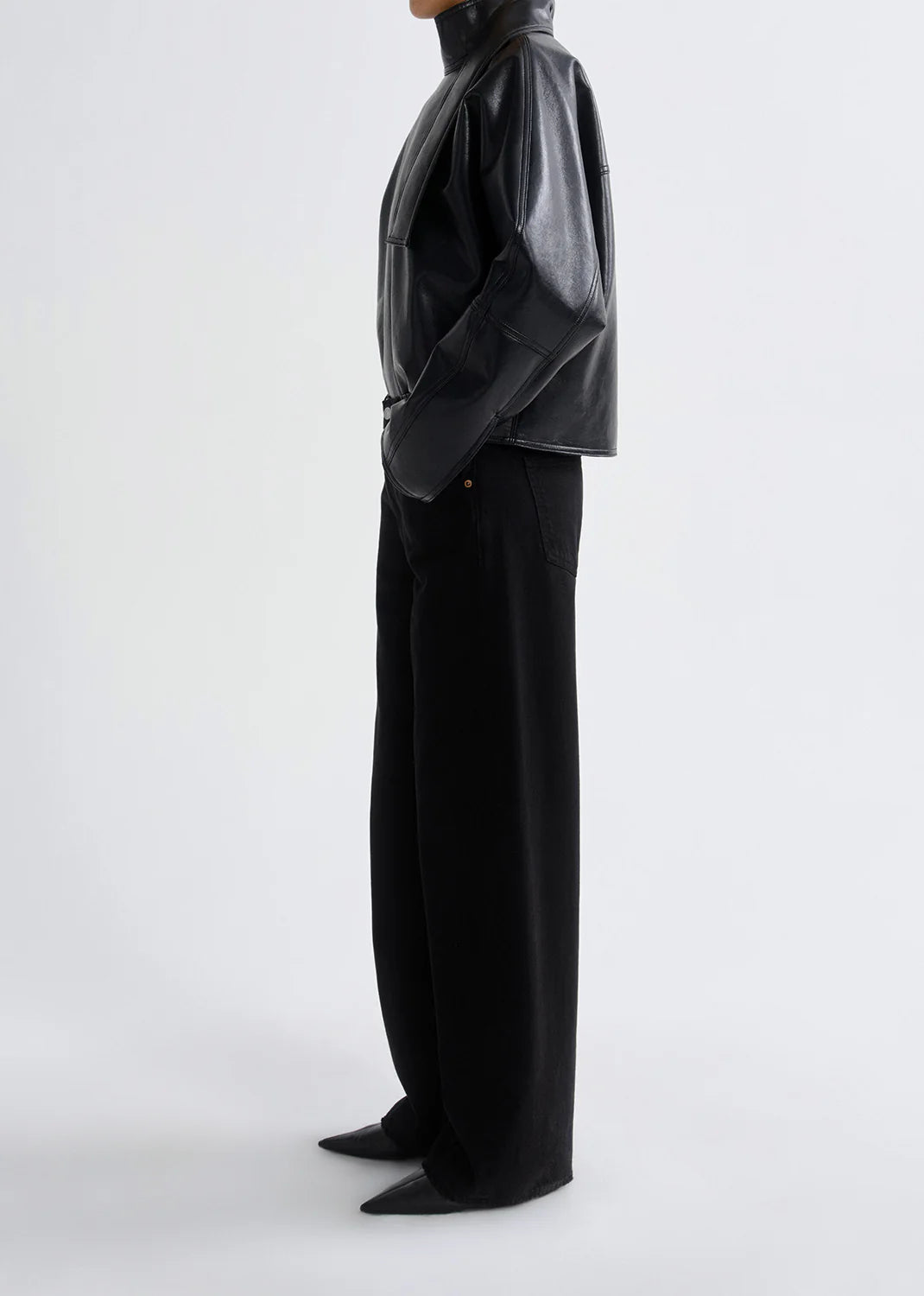 A person stands in profile wearing a black leather jacket, AGOLDE Low Curve Jean - Black, and pointed black shoes against a plain light background.