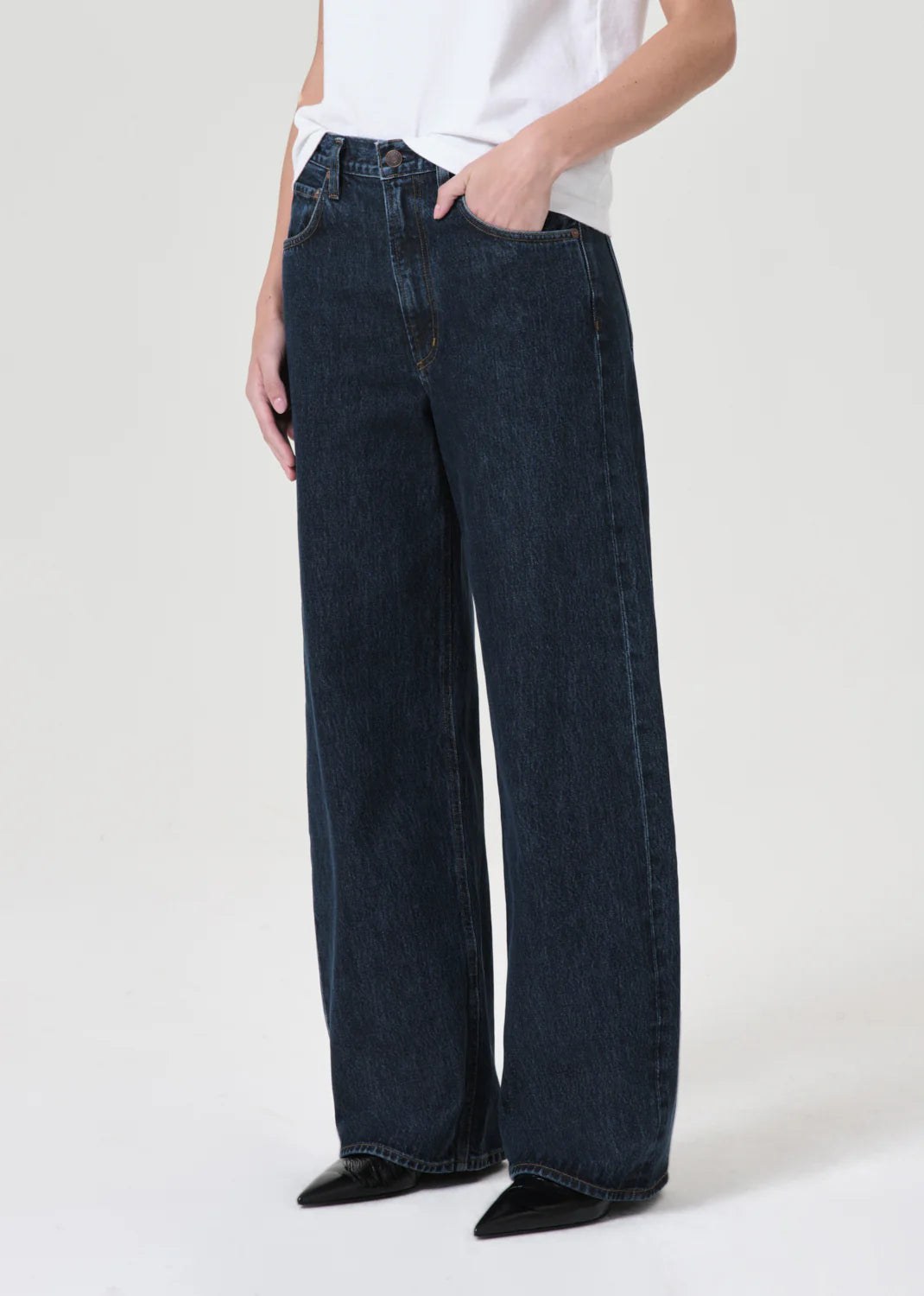A person wearing AGOLDE’s Low Curve Jean - Pendulum in dark blue regenerative cotton and a white t-shirt, with one hand in the pocket, stands against a plain light background.