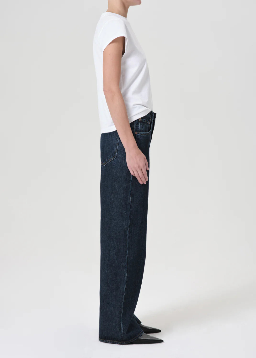 A person stands in profile wearing a white short-sleeve t-shirt, AGOLDE Low Curve Jean - Pendulum in dark non-stretch denim with a mid-rise waist, and black pointed-toe shoes against a plain light background.