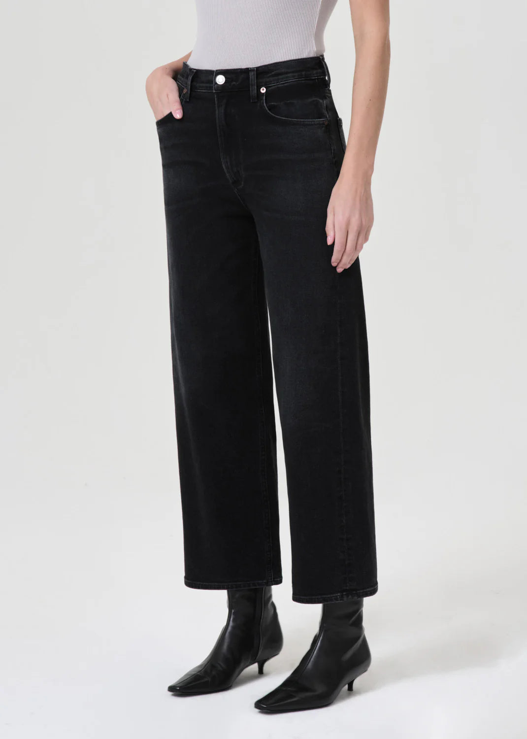 Person wearing AGOLDE Ren High Rise Wide Leg Jeans in Hush, a white top, and black ankle boots. Model has one hand in the pocket and stands against a plain background.
