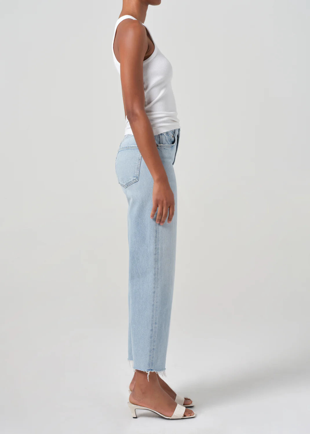 A person in a white tank top, AGOLDE's Ren High Rise Stretch Jeans - Beam made from regenerative cotton, and white heeled sandals stands sideways against a plain background.