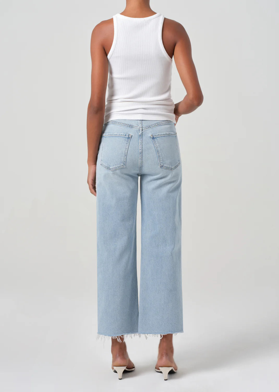 A person with their back turned wears a white sleeveless top, AGOLDE's Ren High Rise Stretch Jeans in Beam, crafted from regenerative cotton, paired with beige heels against a plain background.