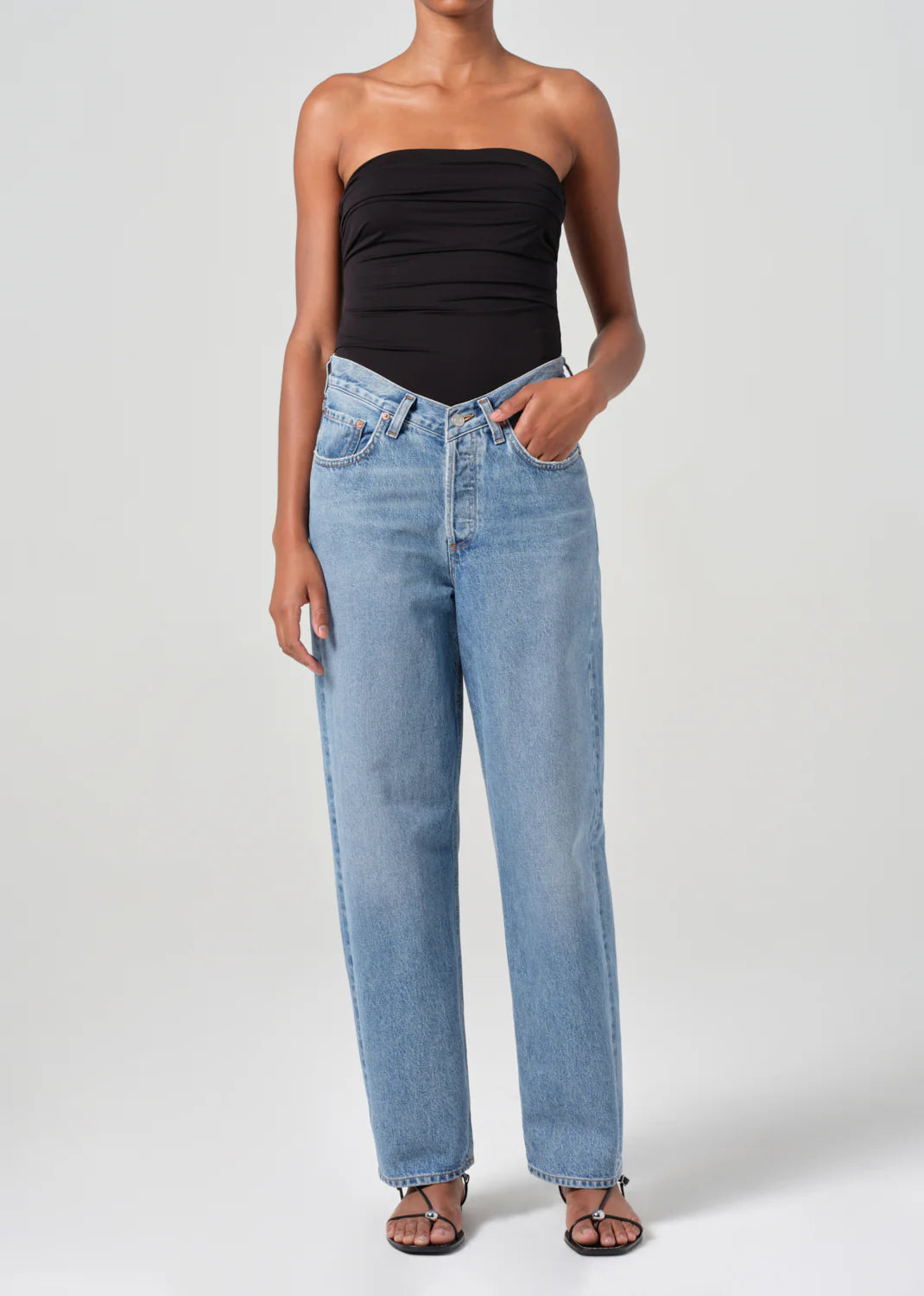 A person wearing a black strapless top and AGOLDE's V-Waist Baggy jeans in Fairway, crafted from regenerative cotton, along with black sandals, stands against a plain background.