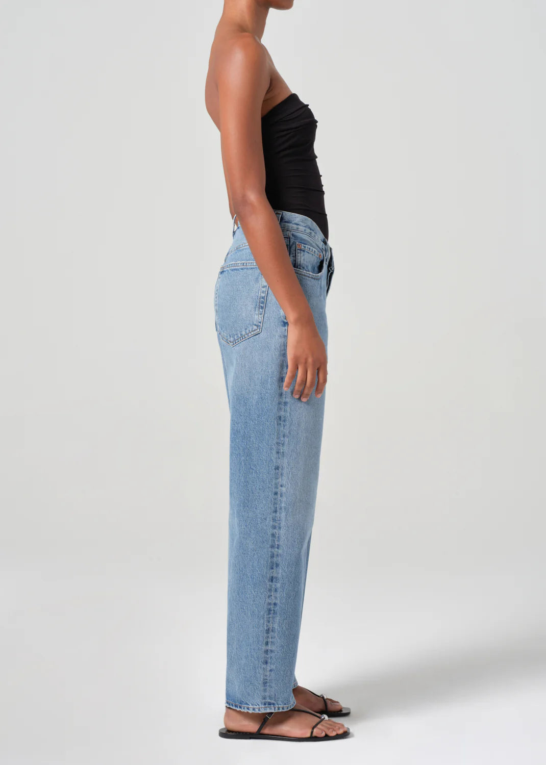 A person stands in profile against a plain background, dressed in a black top, V-Waist Baggy - Fairway jeans from AGOLDE, and black sandals.