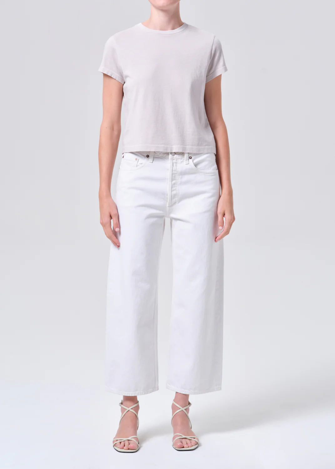 A person stands against a plain light background wearing a short-sleeved light grey t-shirt, AGOLDE Arc Jean 29" - Chalk White high-waisted wide-leg jeans, and white strappy sandals.
