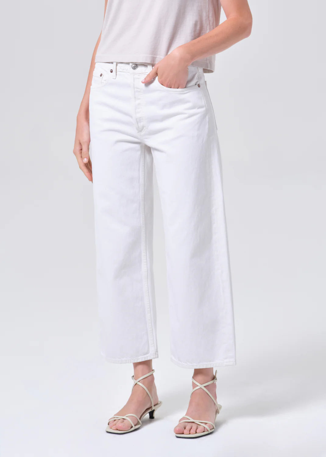 A person wears AGOLDE's Arc Jean 29" in Chalk White—wide-leg, cropped jeans—paired with a light sleeveless top and white strappy sandals, standing against a plain light background.