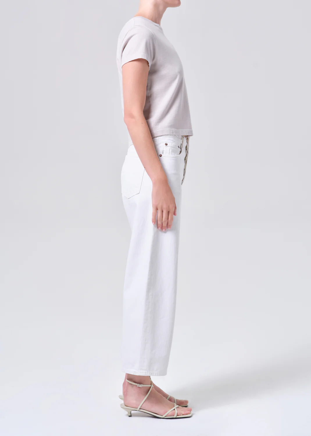 A person stands in profile wearing a light short-sleeve top, flat strappy sandals, and high-waisted AGOLDE Arc Jean 29" in Chalk White made from regenerative cotton, set against a plain background.