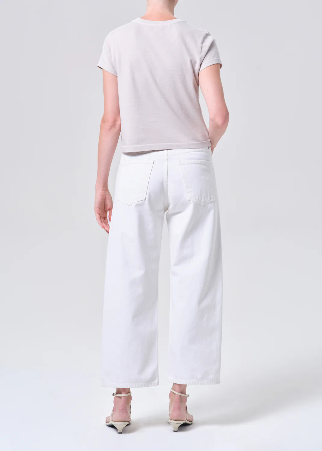 A person stands with their back to the camera, wearing a light short-sleeve shirt, AGOLDE Arc Jean 29" in Chalk White with a relaxed waist and wide leg, paired with white heeled sandals against a plain backdrop.