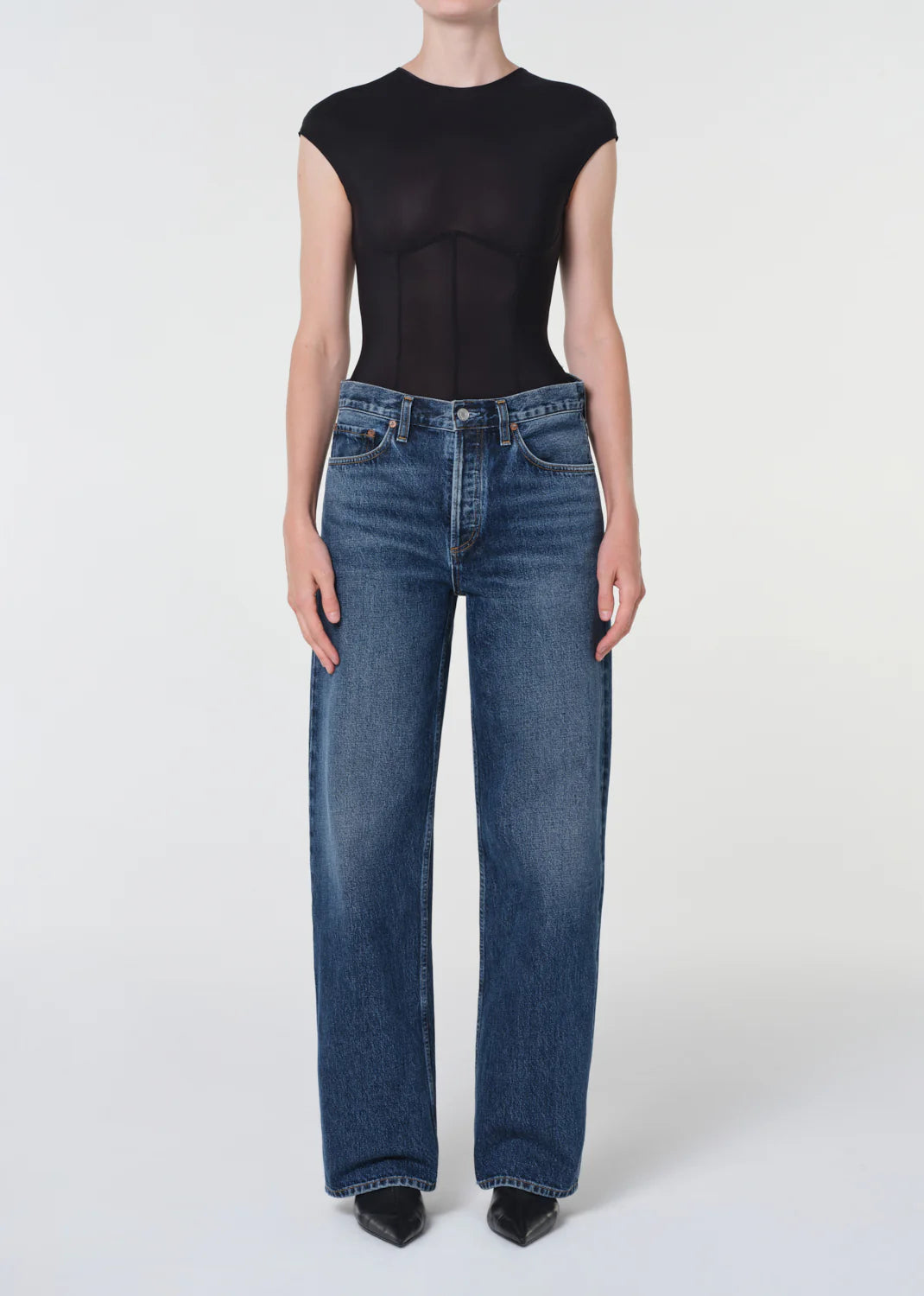 A person in a fitted black short-sleeve top, AGOLDE Arc Jean 32" - Shortwave high-waisted wide-leg jeans, and black pointed shoes stands against a plain white background.