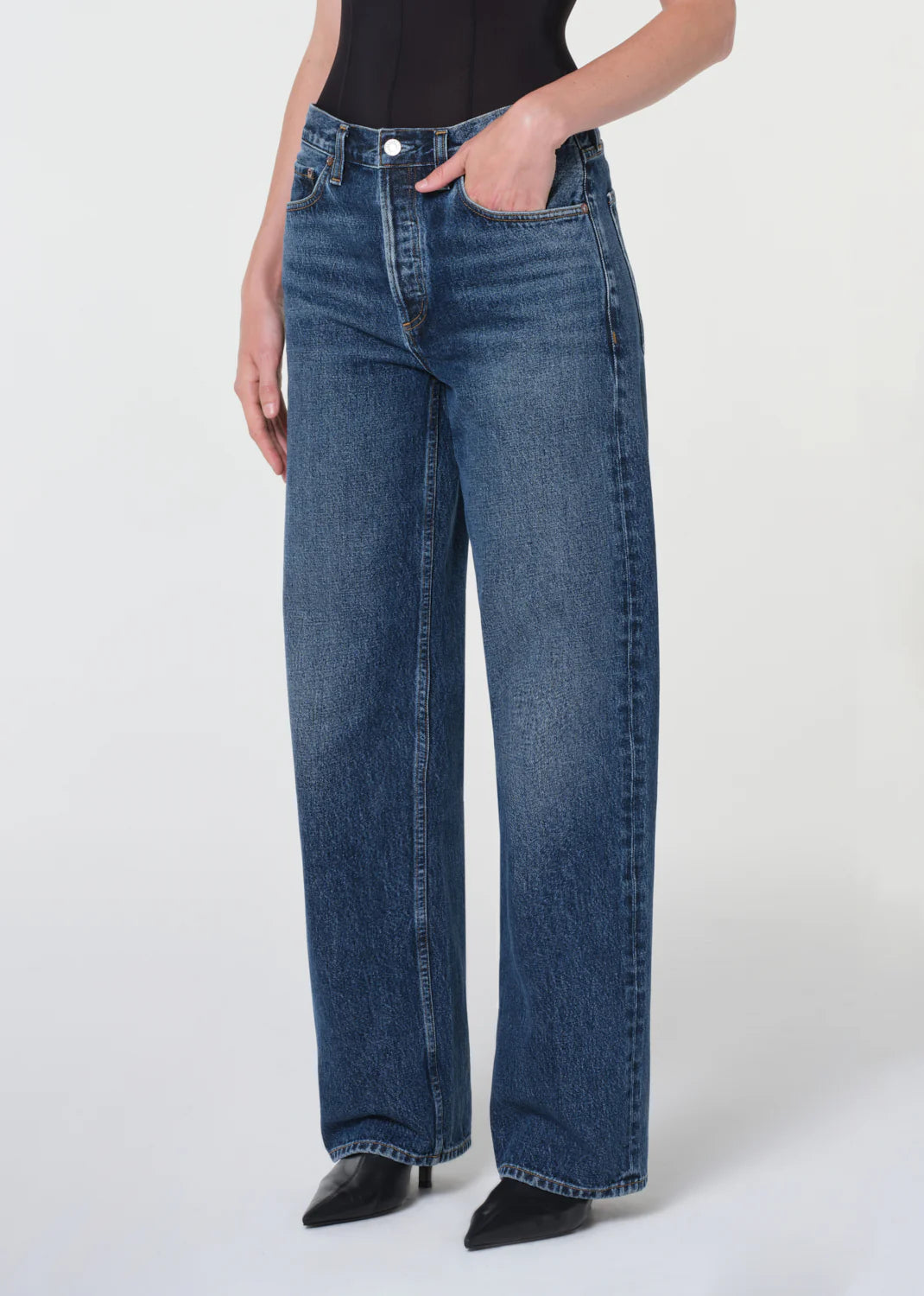 Person shown from shoulders down wearing AGOLDE’s Arc Jean 32" - Shortwave, a high-waisted, wide-leg style in regenerative cotton denim, paired with a black top and black pointed-toe shoes against a plain background.