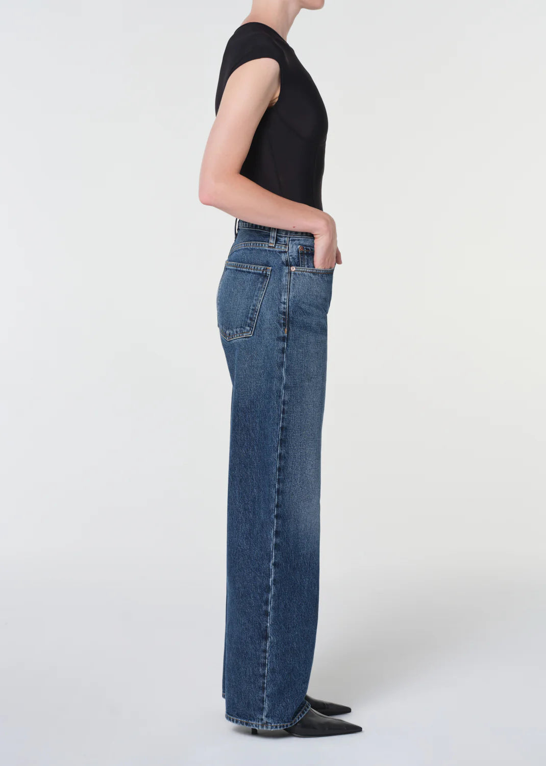 A person in profile wears a black short-sleeve top, AGOLDE Arc Jean 32" - Shortwave high-waisted wide-leg denim jeans, and black pointed-toe shoes against a plain light background.
