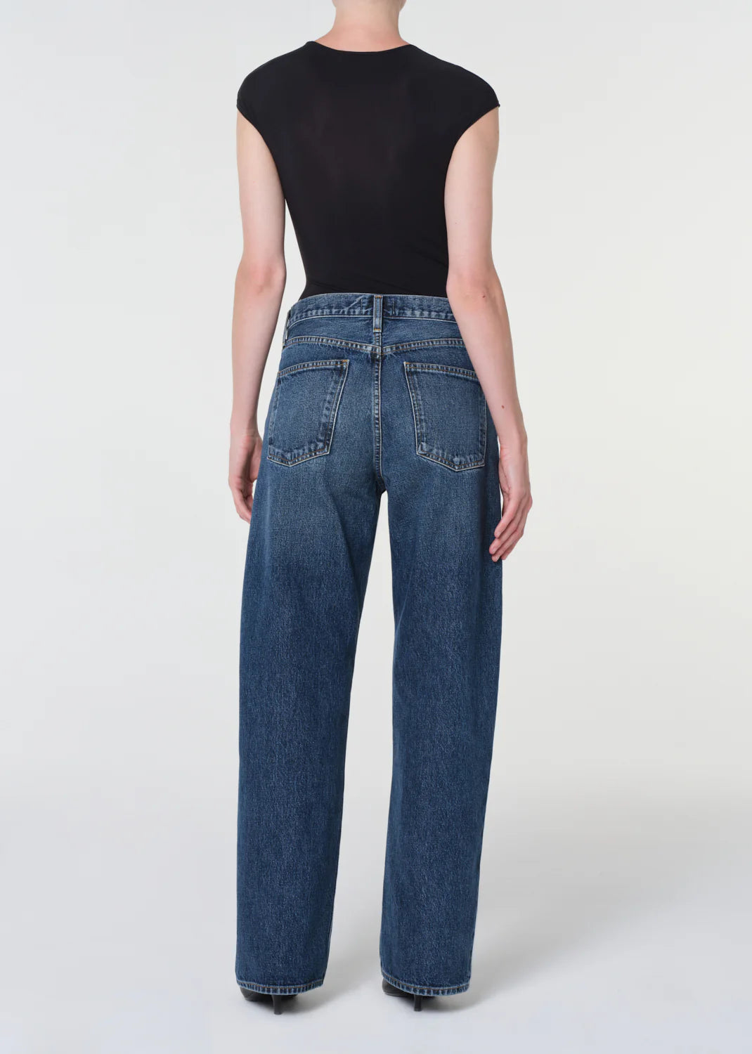 Person stands with back to camera, wearing a black short-sleeve top and AGOLDE Arc Jean 32" - Shortwave in regenerative cotton denim, styled wide-leg against a plain light background.