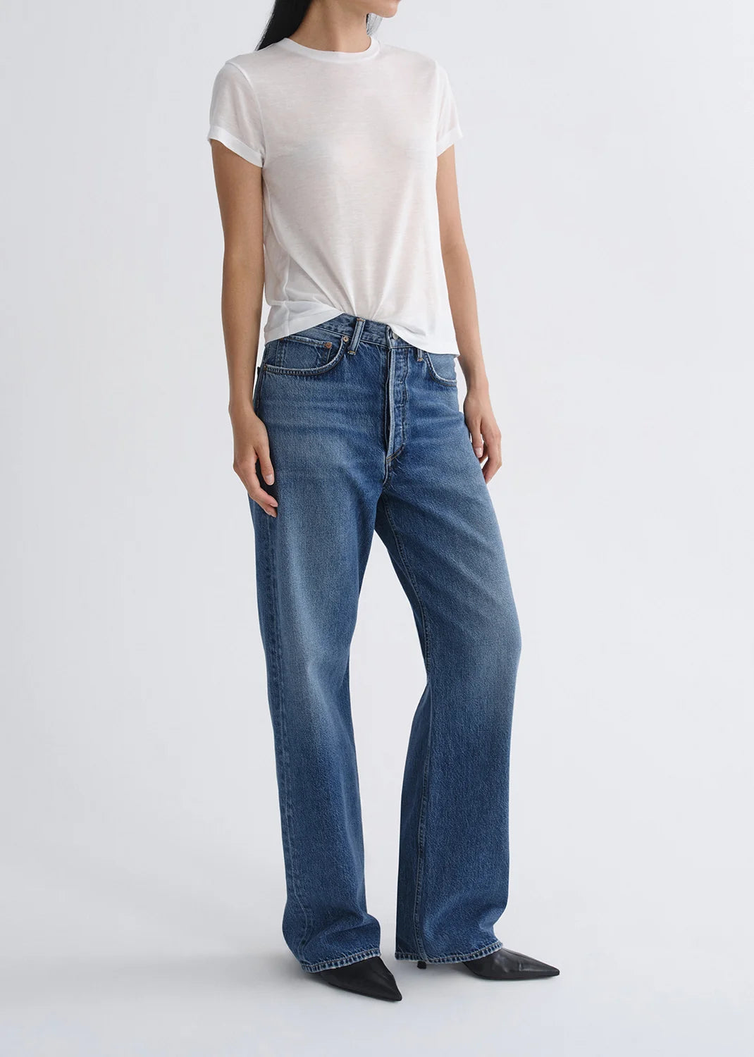 A person wearing a white short-sleeve t-shirt, black pointed-toe shoes, and AGOLDE Vintage Loose - Curb indigo wash straight leg jeans stands against a plain light background.