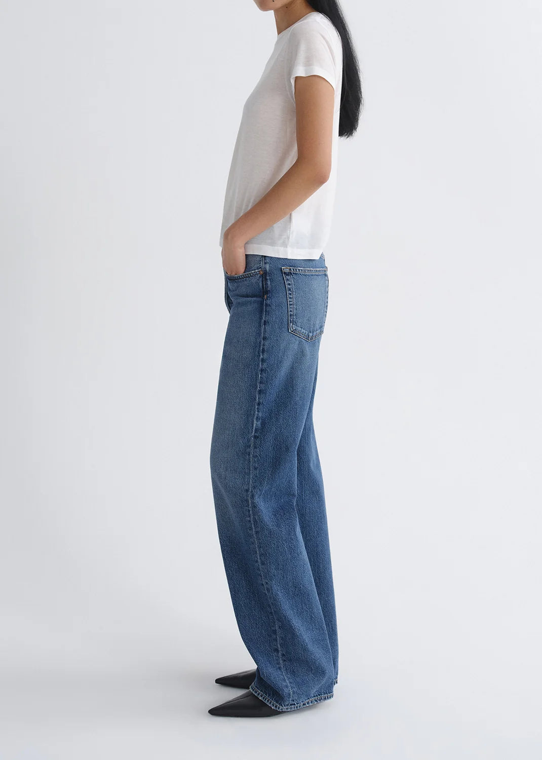 A person standing sideways against a plain white background wears a white short-sleeve t-shirt, black pointed shoes, and AGOLDE Vintage Loose - Curb indigo wash straight leg jeans.