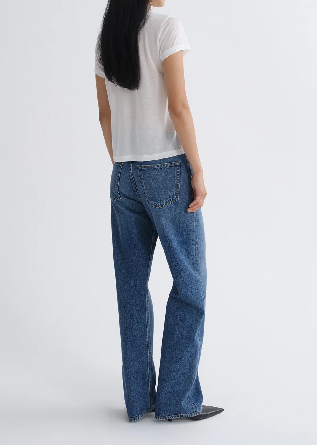 A person stands facing away against a plain light background, wearing a white short-sleeve shirt, black pointed shoes, and AGOLDE’s Vintage Loose - Curb indigo wash straight leg jeans.