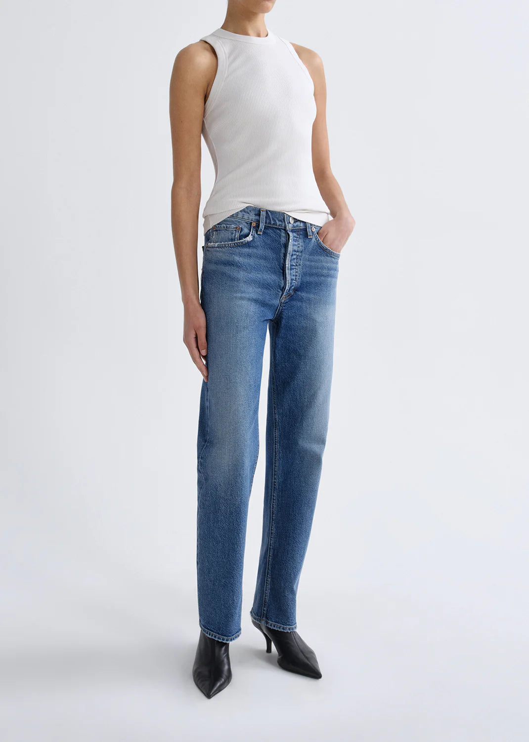 A person stands against a plain background wearing a white sleeveless top, the Lana Jean - Waive (Comfort Stretch) by AGOLDE in a classic straight-leg denim cut, and black pointed-toe shoes.