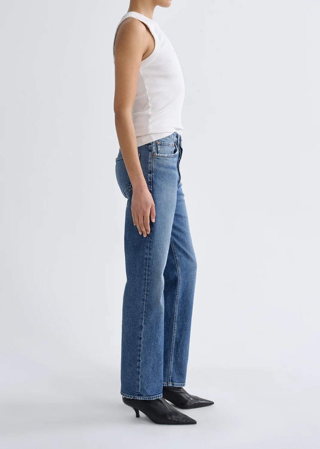 A person stands in profile against a plain background, wearing a sleeveless white top, black heeled shoes, and the AGOLDE Lana Jean - Waive (Comfort Stretch), featuring a vintage indigo wash and a straight-leg denim fit.