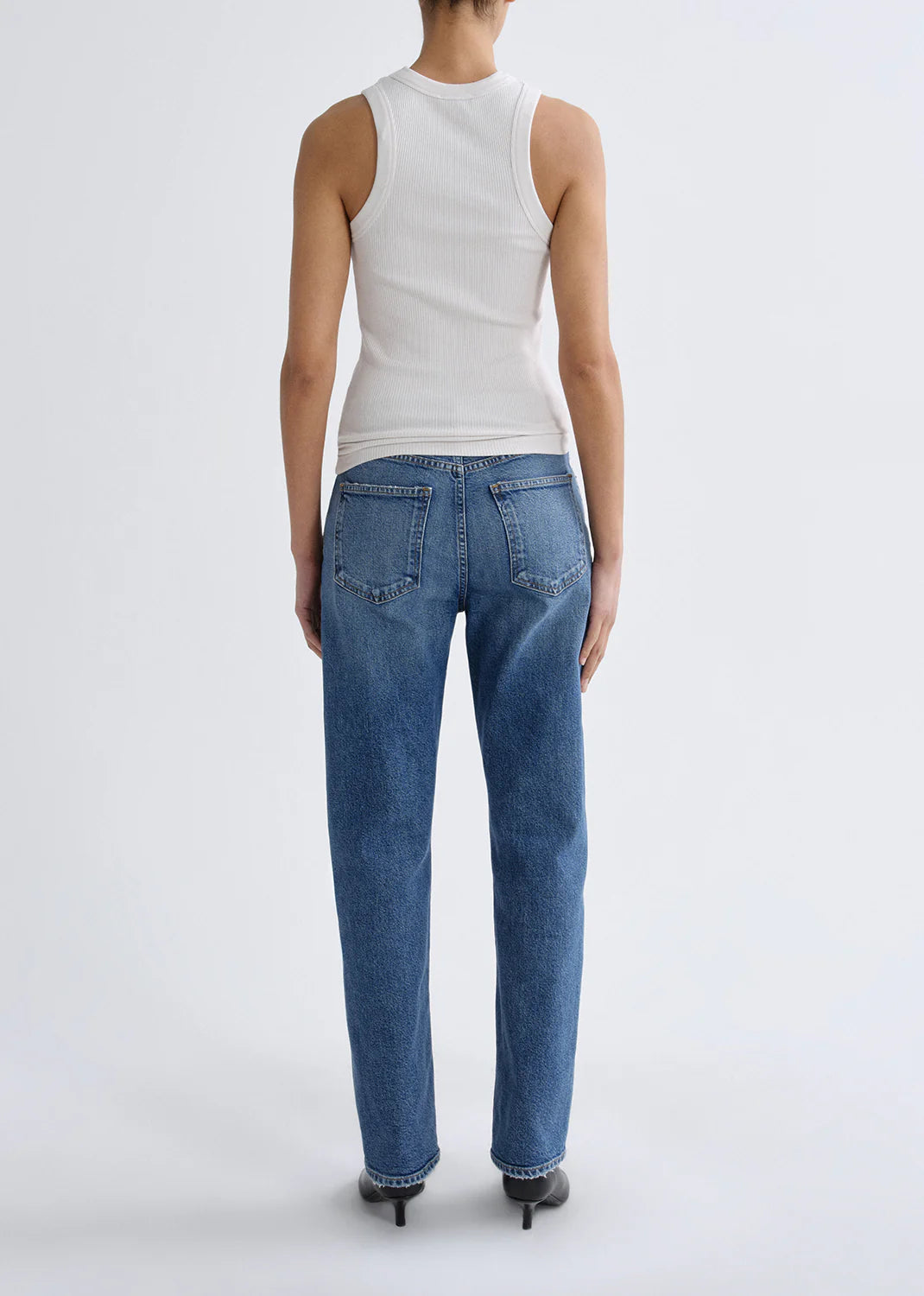 A person stands facing away against a plain light background, wearing a white sleeveless ribbed top, black shoes, and AGOLDE’s Lana Jean - Waive (Comfort Stretch) in vintage indigo.